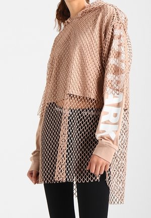 Femme portant un sweat à capuche beige à manches longues avec un texte blanc sur la manche, superposé sous un top en résille beige ample, associé à un pantalon noir.