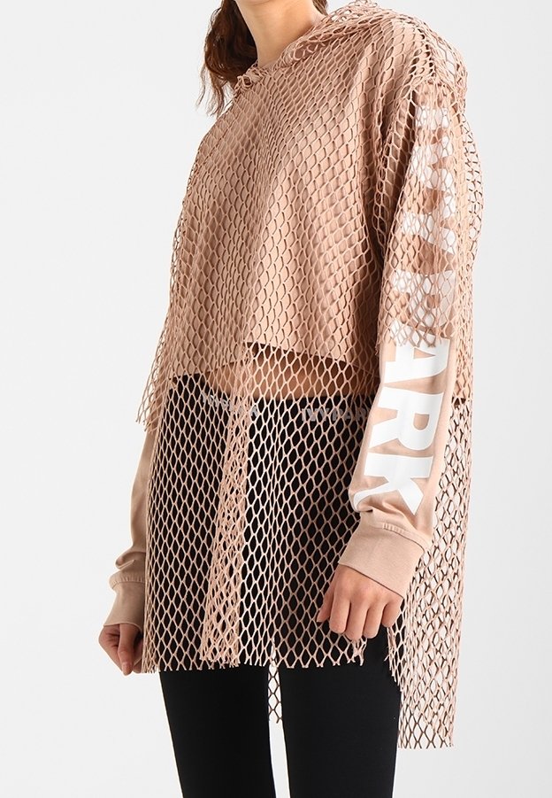 Femme portant un sweat à capuche beige à manches longues avec un texte blanc sur la manche, superposé sous un top en résille beige ample, associé à un pantalon noir.