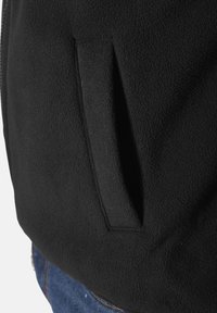 Nahaufnahme einer schwarzen Fleecejacke mit einer seitlichen Tasche im vertikalen Schlitzdesign. Der Stoff hat eine weiche, strukturierte Oberfläche und eine glatte Silhouette.