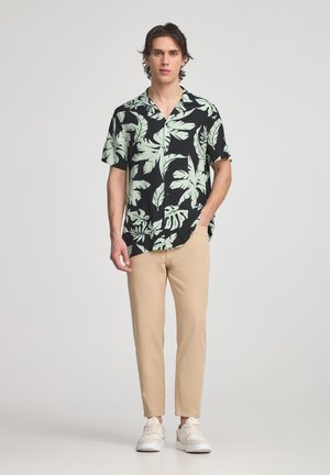 Maglietta nera a maniche corte con stampa di foglie tropicali verdi, abbinata a pantaloni tapered beige e sneakers bianche. Tessuto dalla texture liscia.