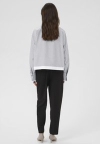 Persoană cu părul lung și brunet, îmbrăcată cu o bluză gri cu mâneci lungi, pantaloni negri și tocuri bej, stând în fața unui fundal alb simplu.