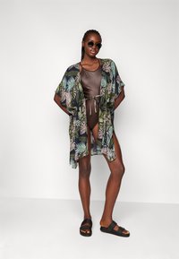 Cubre baño estilo kimono fluido en tela oscura con patrones de hojas tropicales; combinado con un traje de baño marrón y sandalias negras.