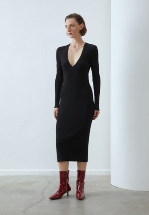 Abito maglione nero a coste con maniche lunghe e scollo a V profondo. Abbinato a stivaletti rossi lucidi alla caviglia con punta affusolata e tacco medio.