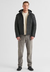 Schwarze Pufferjacke mit Kapuze, gestepptes Design, getragen über einem cremefarbenen Pullover. Helle graue Jeans und schwarze Stiefeletten runden das Outfit ab.