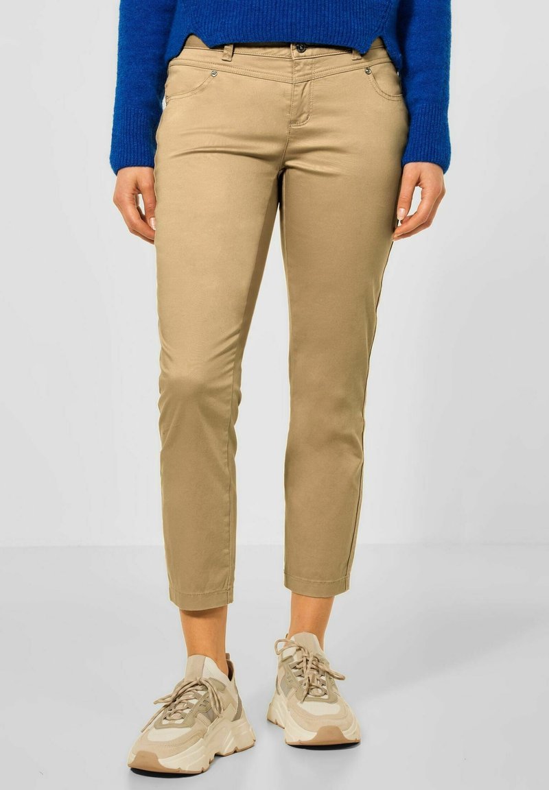 Personne portant un pantalon court beige, des baskets épaisses beiges et blanches et un pull bleu, se tenant devant un fond uni.