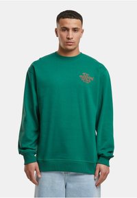 Sudadera verde con cuello redondo, mangas largas y puños acanalados. Presenta un logo impreso de "Do Nothing Club" en marrón en el pecho.