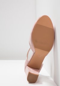 Chaussure à talon bloc en daim rose reposant semelle contre un coin blanc, montrant la semelle en caoutchouc texturée et la bride de cheville.