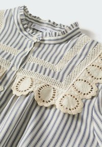 Blouse rayée avec des rayures verticales bleues et blanches, dotée d'une bordure en dentelle avec des découpes circulaires et d'un col froncé. Fermeture à bouton à l'avant.