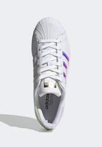 Vit sneaker med en texturerad tåhätta, tre lila och blå ränder, samt en guldlogotyp på tungan. Har ett ventilerande meshfoder.