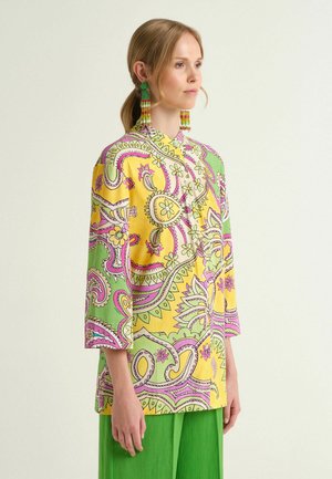 Femme portant un chemisier à manches 3/4 aux motifs jaunes, verts et roses, un pantalon vert et de longues boucles d'oreilles perlées, regardant vers la droite sur un fond blanc.