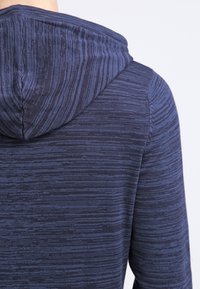 Marineblaue, strukturierte Kapuzenjacke mit Kapuze; verfügt über ein dezentes Streifenmuster und eine körpernahe Passform. Der Stoff wirkt weich und leicht.