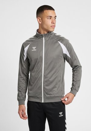 Ung mand iført en grå og hvid sportjakke med lynlås og sorte bukser med hvide hummel-logoer, ser mod højre og justerer jakken med den ene hånd.