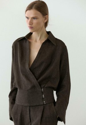 Frau mit hellbraunem Haar, die eine dunkelbraune Wickeljacke mit weitem Kragen und Hakenverschluss trägt, kombiniert mit passenden Hosen.