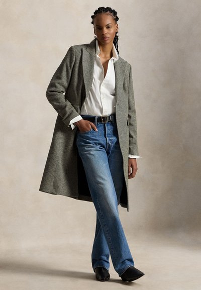 Femme portant un long manteau gris, une chemise blanche boutonnée, un jean bleu, des bottes noires, avec une main dans la poche, debout devant un fond neutre.