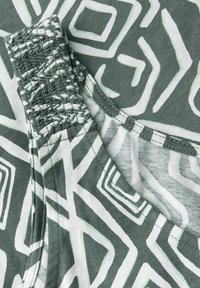 Grön och vit mönstrad tyg med geometriska former, med en texturerad smockad halsringning och släta kanter.