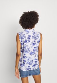 Nainen, jolla on kiharat hiukset, pukeutunut hihattomaan valkoiseen paitapuseroon, jossa on sininen trooppinen kuosi, ja farkkujen minihameeseen, seisomassa yksiväristä taustaa vasten.