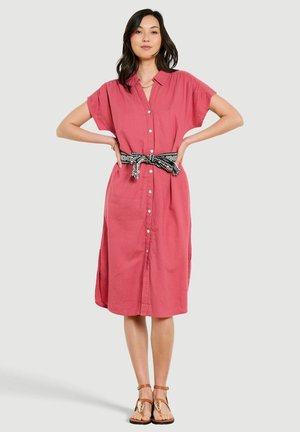 Žena v koralových šatách po kolená zapínaných na gombíky, s krátkymi rukávmi, čiernobielym vzorovaným opaskom a hnedými plochými sandálmi.