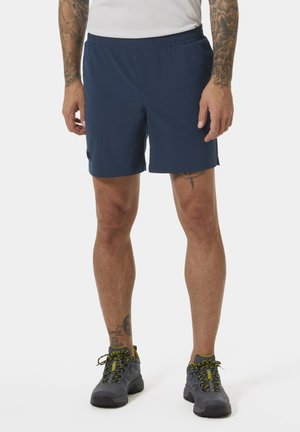 Man draagt marineblauwe sportshort, grijze en neon groene wandelschoenen, en een wit shirt, staand tegen een effen achtergrond.