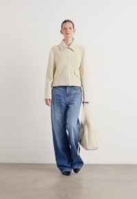 Beige textuur ritsjack, wijduitlopende blauwe jeans, houdt een lichtgekleurde tote tas vast. Eenvoudig ontwerp, neutraal kleurenpalet en ontspannen pasvorm.