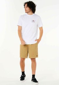 Hvid t-shirt med farverigt grafisk print på brystet, kombineret med khaki shorts i textureret stof og sorte sneakers.