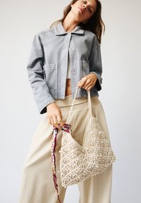 Lichtgrijze spijkerjas met een kraag, gecombineerd met beige wijde broek en een crèmekleurige gehaakte tas met een textuurpatroon en kralenaccenten.