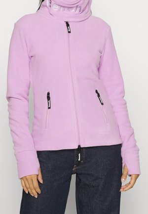 Personne portant une veste polaire violet clair zippée avec des ouvertures pour les pouces et un jean bleu foncé, debout devant un fond uni.