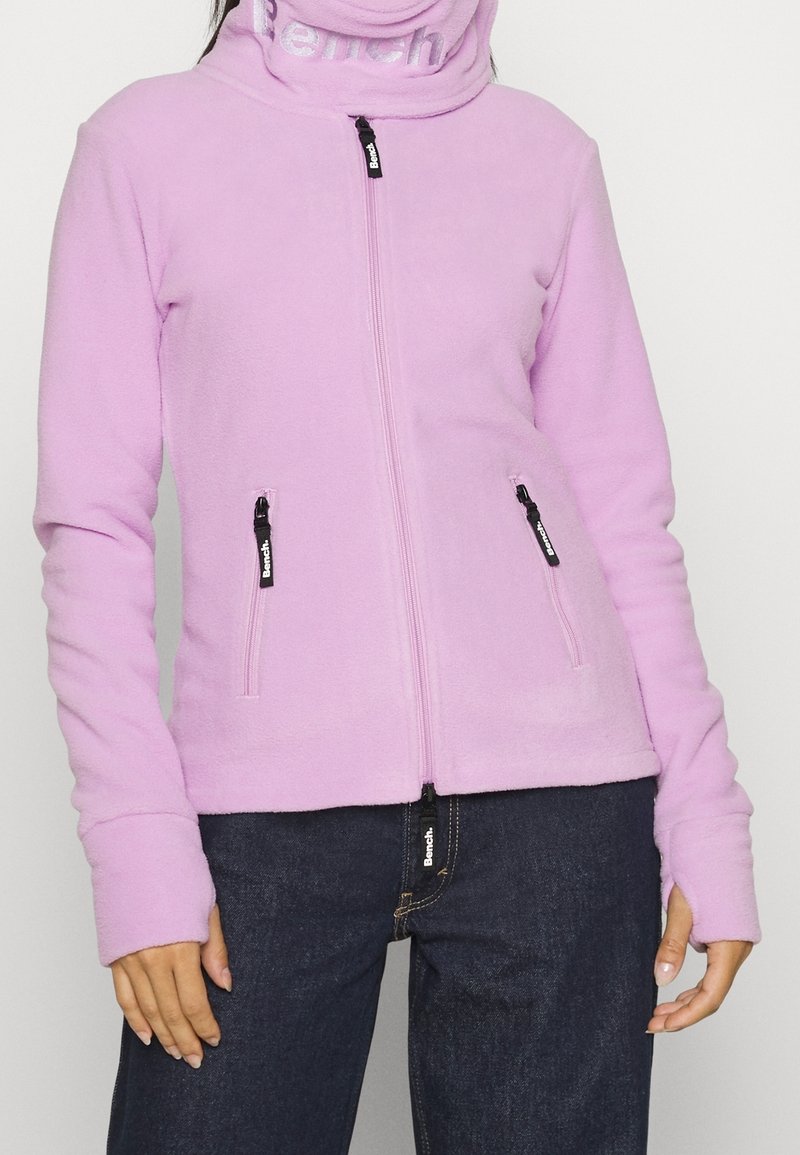 Personne portant une veste polaire violet clair zippée avec des ouvertures pour les pouces et un jean bleu foncé, debout devant un fond uni.