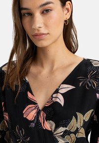 Black dress with floral patterns in pink, cream, and green. Features a v-neckline and textured fabric. Gold hoop earrings add detail.