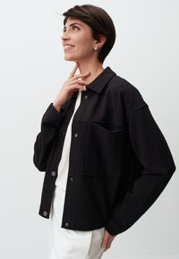 Zwart colbert in een zachte stof met een voorzak, zilveren drukknoopjes en een relaxte pasvorm, gecombineerd met een wit shirt eronder.