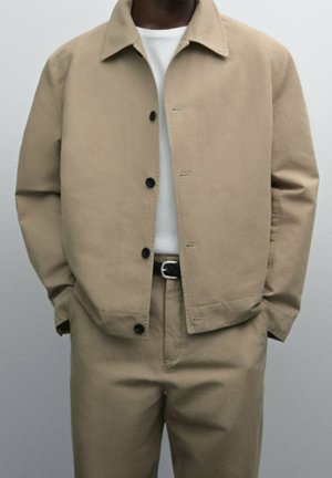 Homme portant une veste beige boutonnée et un pantalon assorti, chemise blanche en dessous, mains dans les poches, debout devant un fond uni.