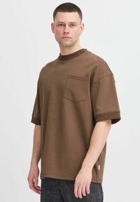 Brun oversized t-skjorte med ribbestrikket hals, korte ermer og brystlomme. Glatt stofftekstur, rett hem og detalj med sideskilt.