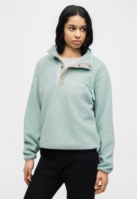 Ανοιχτό πράσινο φλις με επάνω φερμουάρ στο λαιμό, διακοσμημένο με μπεζ λεπτομέρεια και τσέπη με φερμουάρ, υφή επιφάνειας και άνετη εφαρμογή.