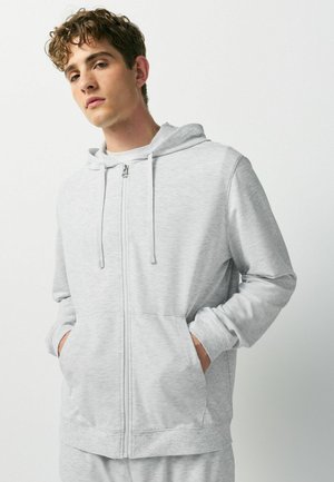 Young man with curly hair wearing a light gray zip-up hoodie with hands in front pockets against a plain background.