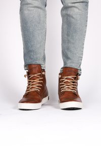 Zapatillas de deporte altas de cuero marrón con cordones beige y suelas de goma blanca. Superficie texturizada y nombre de la marca visible en la lengüeta.