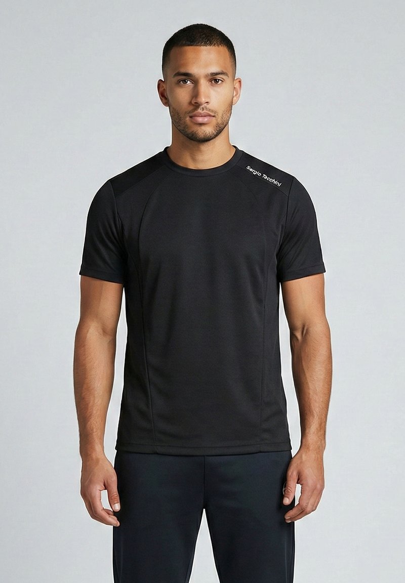 Mees seisab vastamisi, kandes musta lühikeste varrukatega sportlikku särki, millel on vasakul õlal väike valge logo, ning musti pükse.