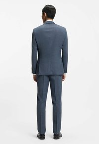 Herrskräddad kostym i blå texturerad tyg; figursydd kavaj med notslag, byxor med raka ben och eleganta svarta skor. Bakre vy.