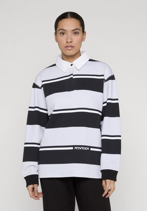 Donna con capelli scuri che indossa una maglia da rugby a larghe righe bianche e nere con colletto bianco e logo "PEGADOR" sul davanti.