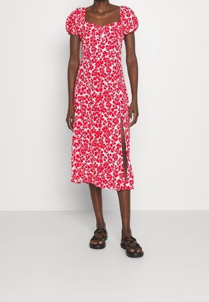 Kobieta ubrana w czerwono-białą sukienkę midi w kwiaty z bufkami i rozcięciem sięgającym uda, w połączeniu z czarnymi sandałami na paski.