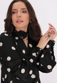 Blusa nera con grandi pois beige, presenta un colletto a forma di rosa, bottoni con accenti di perla e una texture liscia simile alla seta.