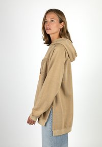 Beige Kapuzenpullover mit großer Kängurutasche und seitlichen Schlitzen. Hergestellt aus weichem Stoff, mit einem gestickten Logo in Orange auf der Vorderseite.