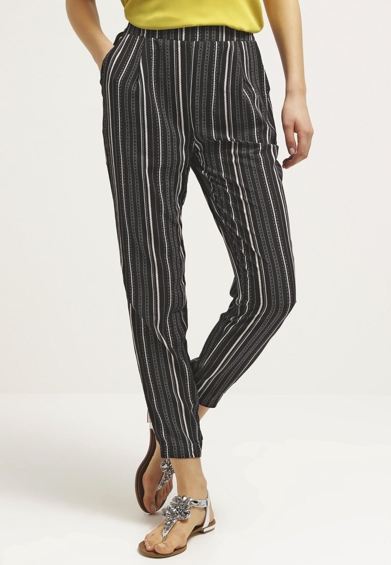 Femme portant un pantalon noir à rayures blanches verticales, un haut jaune et des sandales argentées ornées de bijoux, debout avec une main dans la poche.