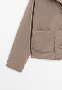 Short brown jacket made of lightweight fabric, featuring a rectangular pocket and round buttons, with a cropped design and smooth texture.