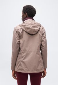 Άτομο με ανοι χτό καφετί αδιάβροχο με κουκούλα και βυσσινί παντελόνι, στέκεται με την πλάτη προς τον θεατή σε απλό φόντο.