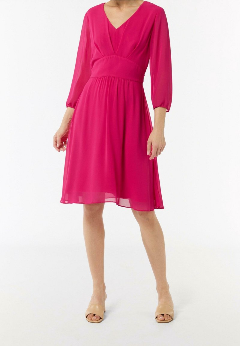 Femme portant une robe rose mi-longue aux manches trois-quarts et des talons nus à bout ouvert, debout sur un fond blanc.