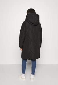 Svart vadderad jacka med huva, med en diagonalt quiltad mönster, lång ärm och mittenlängd design. Bärs över blå jeans och ljusa sneakers.