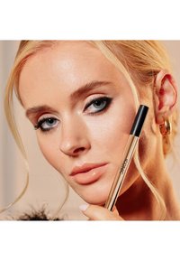 Gull flytende eyeliner penn med svart hette holdt nær ansiktet. Modellen har lys hud, blond hår, og har på seg mørk øyemakeup.