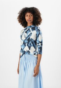 Langsleeve top in blauw en crèmekleurig tie-dye patroon met een hoge hals. Gecombineerd met een lichtblauwe rok met elastische taille. Zachte stoftextuur.