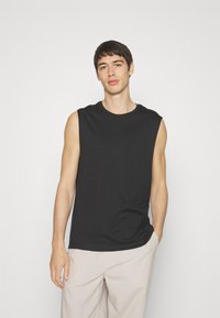 T-shirt nera senza maniche in cotone, con scollatura rotonda e vestibilità rilassata, abbinata a pantaloni chiari.