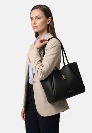 Beigen bleiserin nainen kantaa olallaan suurta mustaa nahkalaukkua vaaleaa yksiväristä taustaa vasten.