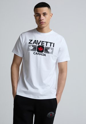 Jeune homme aux cheveux courts portant un T-shirt blanc Zavetti Canada et un pantalon noir, les mains dans les poches, sur un fond gris uni.